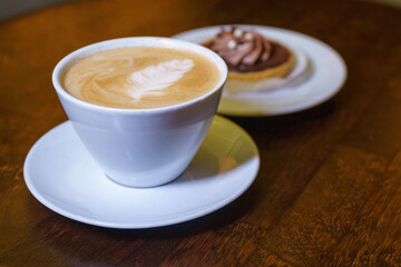 cup of cappuccino with Latte art on top is on the wooden table and cake in soft focus on background