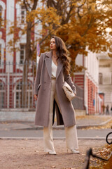 Elegant brunette woman with long wavy hair wearing grey wool coat, holding white handbag and walking at city street on autumn day. Portrait against yellow leaves