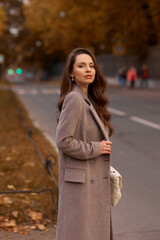 Fototapeta premium Elegant brunette woman with long wavy hair wearing grey wool coat, holding white handbag and walking at city street on autumn day. Portrait against yellow leaves