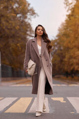 Elegant brunette woman with long wavy hair wearing grey wool coat, holding white handbag and...