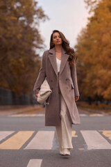 Elegant brunette woman with long wavy hair wearing grey wool coat, holding white handbag and walking at city street on autumn day. Portrait against yellow leaves