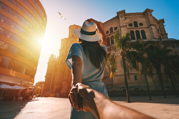 Follow me - POV. Young tourist woman in white sun hat holding her boyfriend by hand and walking in Malaga city at sunset. Couple on summer holiday vacation in Spain. Traveling together. - obrazy, fototapety, plakaty