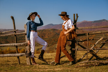 Two fashionable women wearing stylish autumn outfits with trendy hats, sunglasses, bags, cowboy boots, posing in mountains. Copy, empty space for text