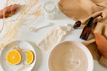 Beauty spa beige composition with amber bottle, bowl with water, orange slices, cotton bag, bath salts on white background. Flat lay, top view. Female beauty treatment routine concept.