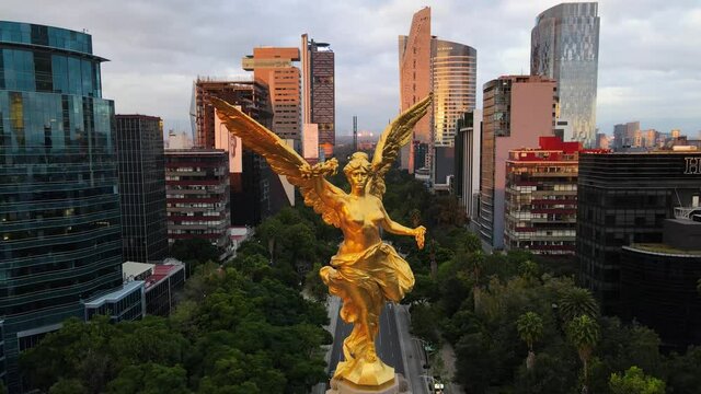 Toma a&eacute;rea del monumento a la glorieta del &aacute;ngel al amanecer con los rascacielos de la ciudad de M&eacute;xico de fondo haciendo un paneo hacia abajo para revelar la base adornada de flor de cempas&uacute;chil 