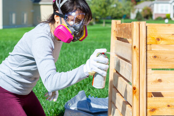 A young caucasian woman wearing face mask safety goggles and gloves is spray staining  a wooden...