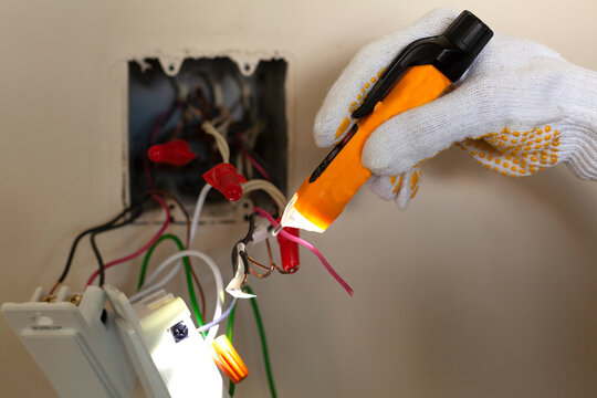 An electrician is replacing a wall switch. A DIY project concept. High voltage danger. A touchless voltage tester and protective rubber gloves are used. The tester has torch light for illumination