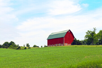 Obraz premium bright red farm barn building on a bright green pasture hillside under cloudy blue sky