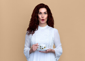 Woman with Fun Surprised Look Holding Vintage Teacup on Beige Background. Studio portrait of woman...