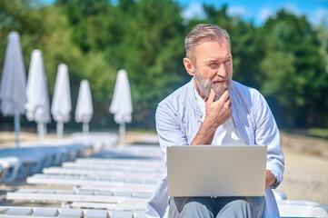 Pensive man with laptop sitting on beach lounger