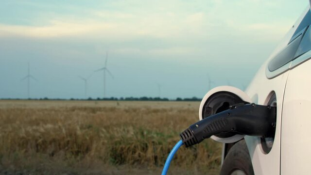 Wind driven generator charges electric Tesla. Powerful wind turbine farm in golden ripe wheat field and ev charged at station on foreground