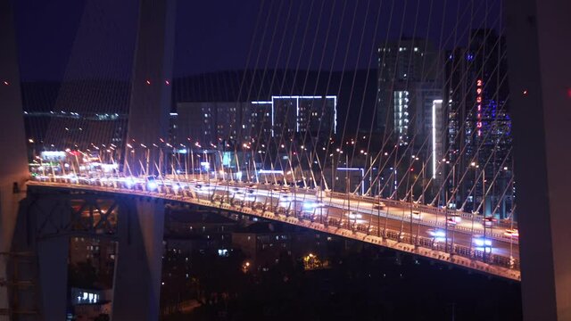Great timelapse of the cable-stayed Golden Bridge across the Zolotoy Rog (Golden Horn Bay) and many busy cars driving along the road. Night. Vladivostok cityscape is on the background