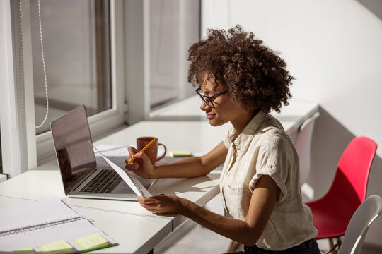 Happy pretty woman wearing glasses and sitting at workplace while holding document and reading