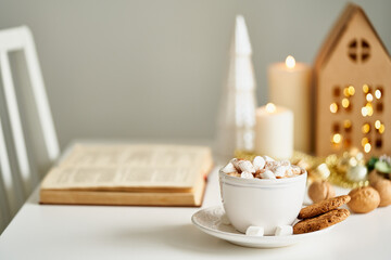 Reading book on cozy winter evening with candles. Christmas light gray background with copy space. Cocoa hot chocolate with marshmallows and cookies. Side view of table, chair in home room