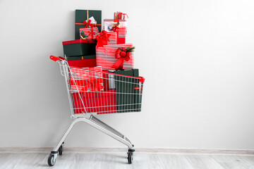 Shopping cart with beautiful Christmas gifts near light wall