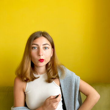 Funny caucasian young woman with bright red lips. Girl twists her mouth thinking of something and pushes the remote button. Yellow background. Picture with copyspace