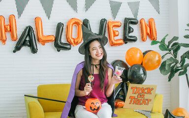 Obraz na płótnie Canvas Portrait Asian beautiful woman wearing witch costume for decorating and celebrating Halloween party, smiling with happiness, holding candy and lollipop while sitting in living room at home.