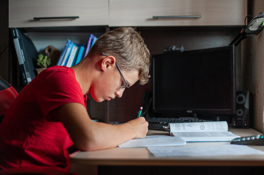 A teenage boy in a red T-shirt sits at a table at home and does his homework. A boy with glasses writes in a notebook and looks at a textbook. Study without gadgets.