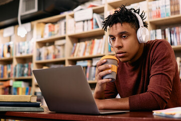 Young black student drinking coffee while e-learning on laptop in a library.
