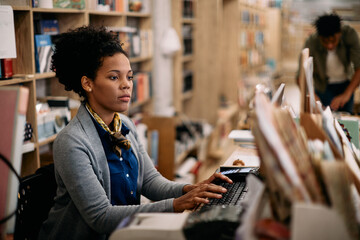 African American woman using desktop PC while working at bookstore. - obrazy, fototapety, plakaty