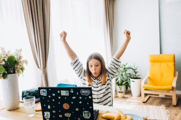 a girl of 11 years old plays computer games on a laptop, rejoices, raises her hands up