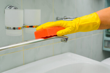 Household cleaning, bathroom washing. Close-up of female hand in rubber glove with sponge wiping chrome faucet. Side view of housewife cleaning from dirt and rust indoors