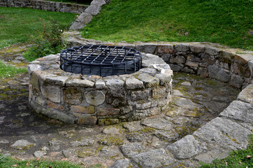 stone round fountain with a protective grid of twisted prisms. forged steel bars go spiraled at the top. around the well there is a retaining wall and a lawn. protection against falling into the depth