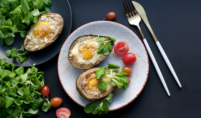 Baked avocado with egg, cheese, corn salad and tomatoes on a white plate on black background. Breakfast with avocado. Gold knife and fork. Top view