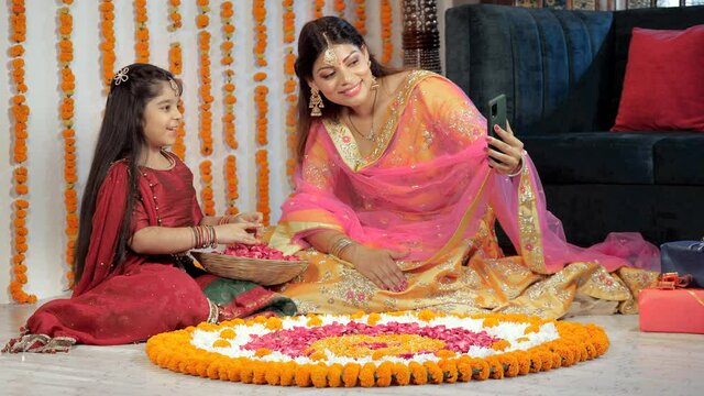 A young mother and her sweet child talking on a video call in the festive season. Pretty female and her daughter in ethnic wear sitting together against the floral background - Hindu festival  Diwa...