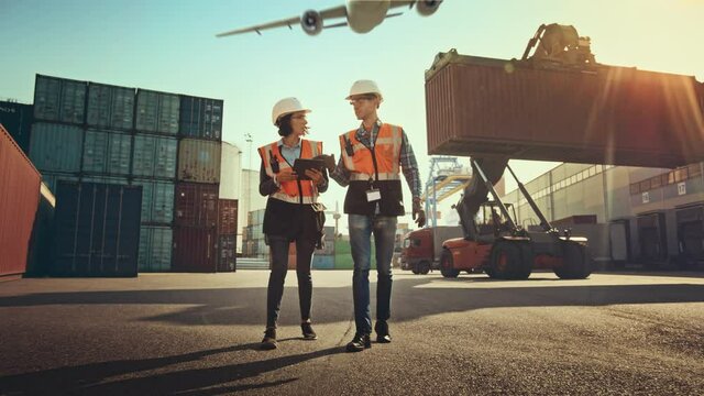 Female Industrial Engineer with Tablet Computer and Male Foreman Worker in Hard Hats Walk in Container Terminal. VFX Airplane Fly in the Sky and Double Girder Gantry Cranes Work in the Background.