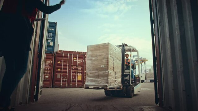 Forklift Driver Loading a Shipping Container with a Pallet with Boxes in Logistics Terminal. Female Industrial Supervisor Helping the Process. VFX Double Girder Gantry Cranes Work in the Background.