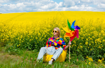Obraz na płótnie Canvas Beautiful female in 90s stylish shirt with pinwheel sitting in rapeseed field
