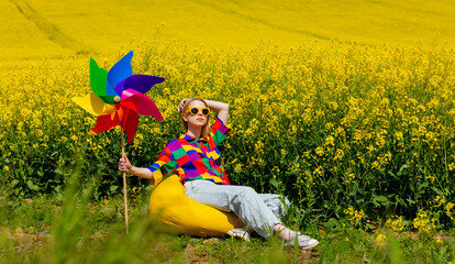 Beautiful female in 90s stylish shirt with pinwheel sitting in rapeseed field