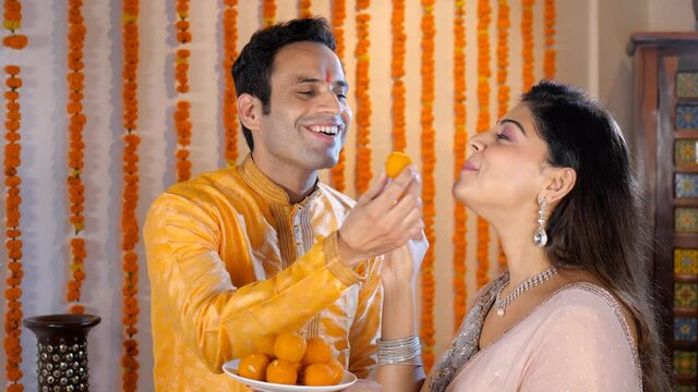 Middle-aged husband-wife feeding sweets to each other on a festive occasion. An Indian man and his wife standing against a floral background- Diwali decorations  a Hindu festival  an auspicious occ...