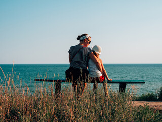 Happy mother and daughter sitting on bench on marine landscape back view. Mom and child having fun...
