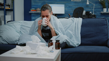 Adult with seasonal flu bowing runny nose with tissue and drinking cup of tea against virus infection. Woman feeling cold wrapped in blanket waiting for treatment to cure sickness.