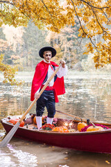 Obraz premium young man dressed as gondolier in gondola boat decorated with pumpkins on pond in autumn park celebrates Halloween and having fun, concept of Halloween carnival or costume party