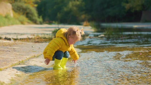 A happy little girl in yellow rubber boots stand in river and plays with the water. Slow motion.
