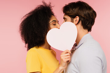 interracial couple with closed eyes kissing behind blank paper heart isolated on pink.