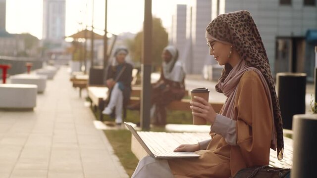 Medium long side view of young Middle Eastern woman wearing hijab and eyeglasses, sitting on bench on pedestrianized street, video calling via laptop computer