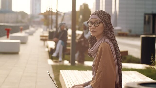 Medium long side view of young Middle Eastern woman wearing hijab and eyeglasses, sitting on bench on pedestrianized street, holding portable computer on lap, looking on camera