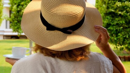 female age 50-55 in a straw hat holds a cup of hot drink and drinks coffee, sitting in the morning on a summer terrace on a sunny day. The concept of good morning, travel and expectation of new