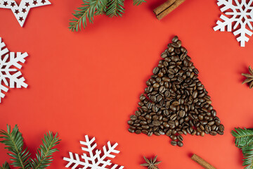 Brown coffee beans laid out in shape of Christmas tree with spruce branches and wooden snowflakes on red background