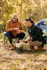 camping, tourism and travel concept - happy couple drinking beer and cooking food in pot on tourist gas burner at tent camp