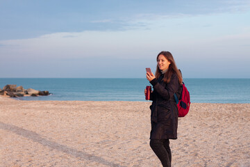 Happy young woman in black raincoat with backpack on empty beach autumn sea. Smiling millennial girl with long hair walking hiking drinking tea in thermo can bottle alone.Lifestyle real people outdoor