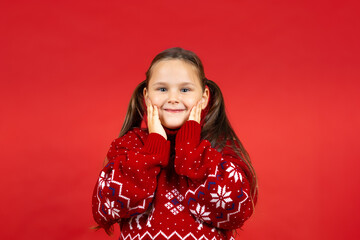 close-up portrait of beautiful happy girl in red knitted Christmas sweater with reindeer touching her face with hands, isolated on red background. 