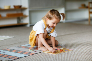 Cute caucasian girl toddler sits and plays on the floor on a rug with a wooden puzzle in a bright...
