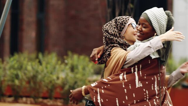 Locked-down of young Muslim Middle Eastern woman standing in park, her Black female friend coming and hugging, friends talking at daytime