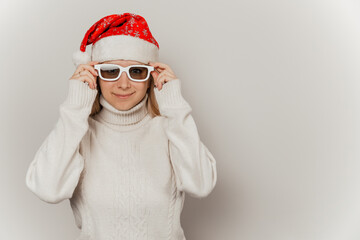 Portrait of cute smiling girl in white sweater, santa hat and glasses on gray isolated background