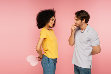 cheerful hispanic woman holding paper heart behind back near curious boyfriend isolated on pink.